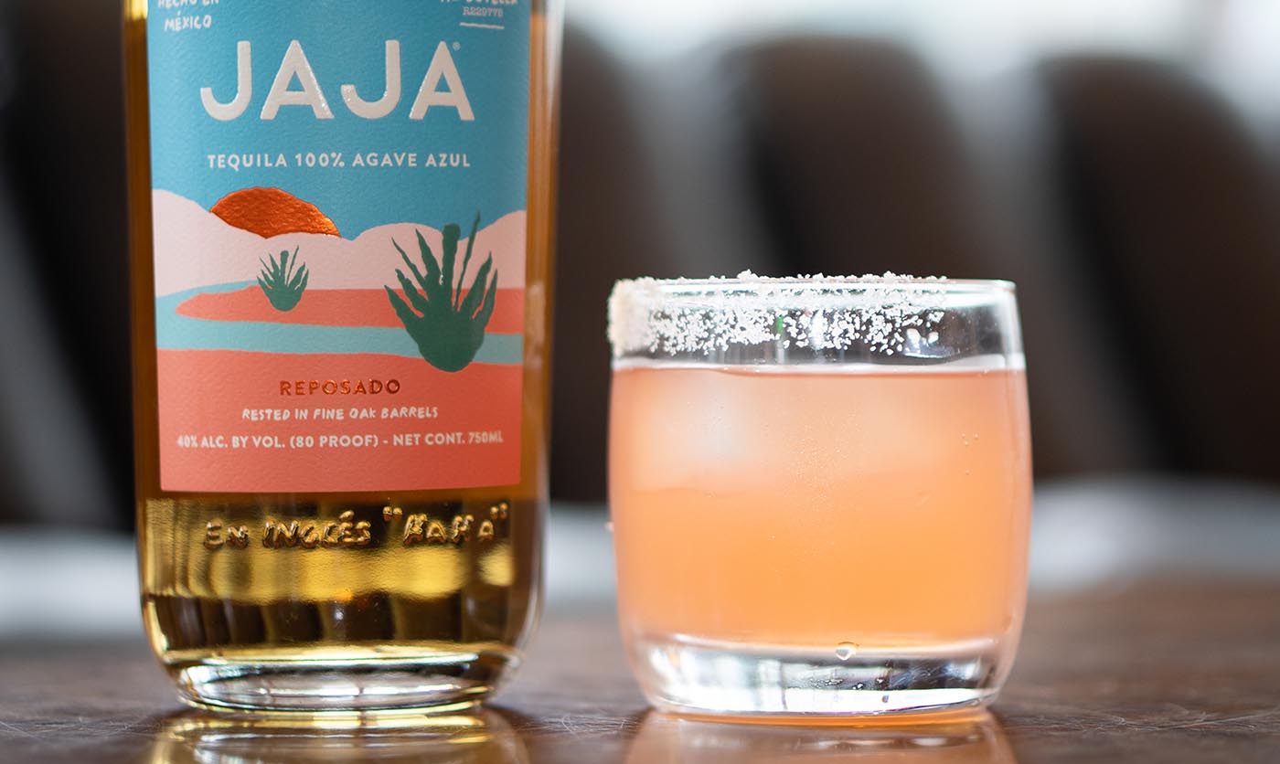 A bottle of reposado JAJA Tequila next to a blood orange puree cocktail in a tumbler.