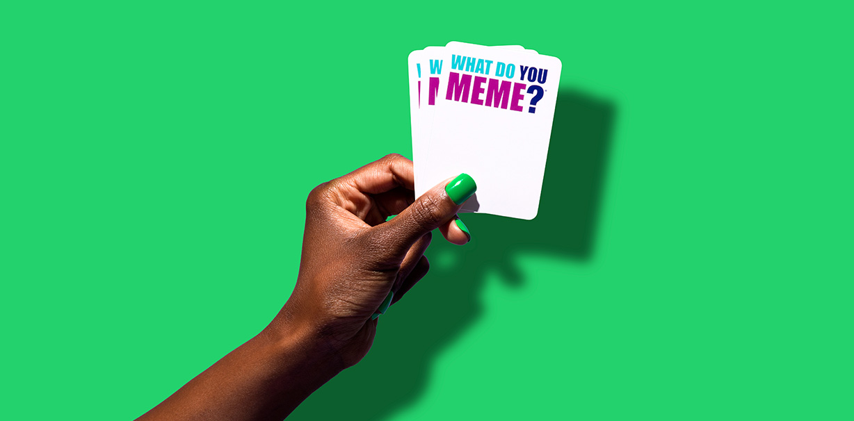 A hand holding What Do You Meme? cards in front of a green background.