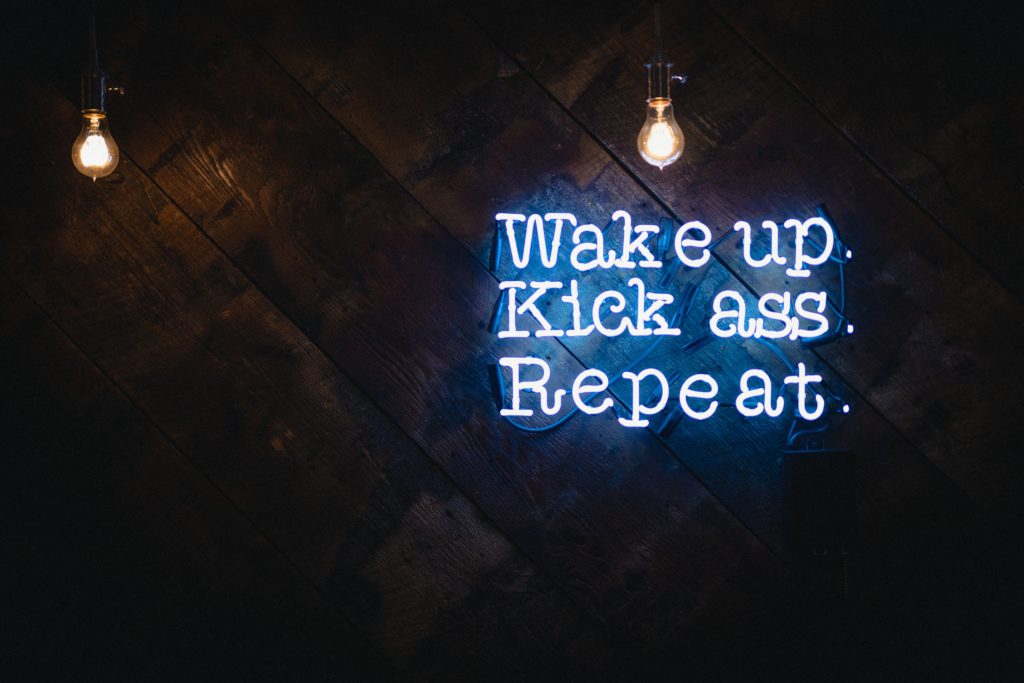 Neon sign on wall of dimly lit room. States “Wake up. Kick Ass. Repeat”